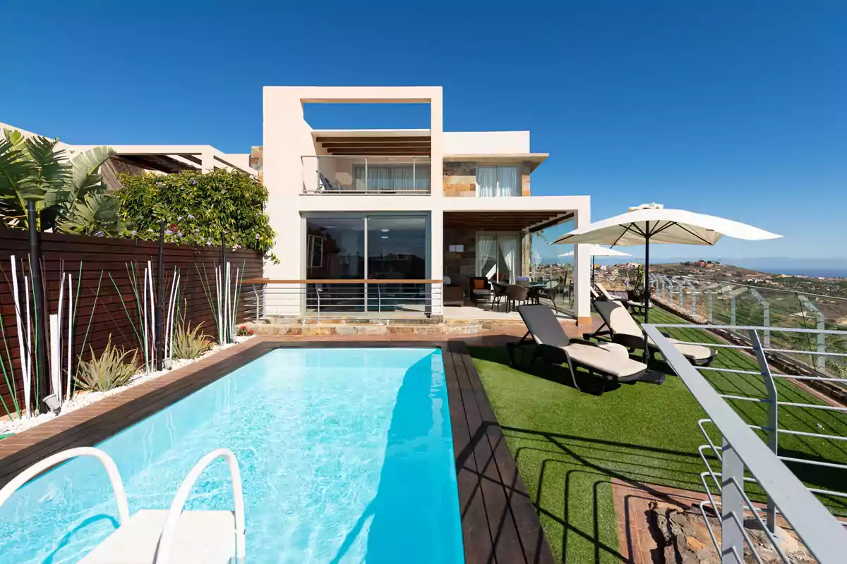 Casa moderna con piscina, tumbonas y sombrilla en un entorno soleado con vista panorámica.