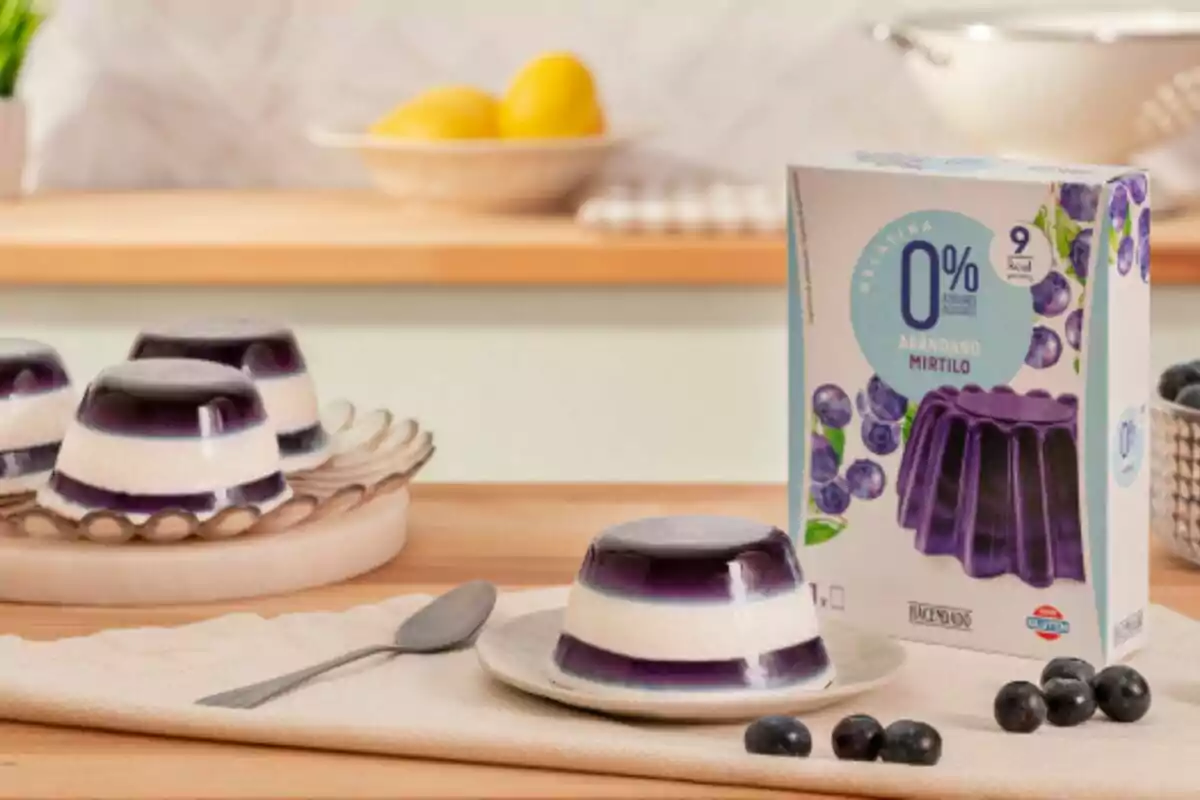Postre de gelatina de arándano y crema en un plato con una caja del producto al lado y arándanos frescos alrededor sobre una mesa de madera. Postre de gelatina de arándano y crema en un plato con una caja del producto al lado y arándanos frescos alrededor sobre una mesa de madera.