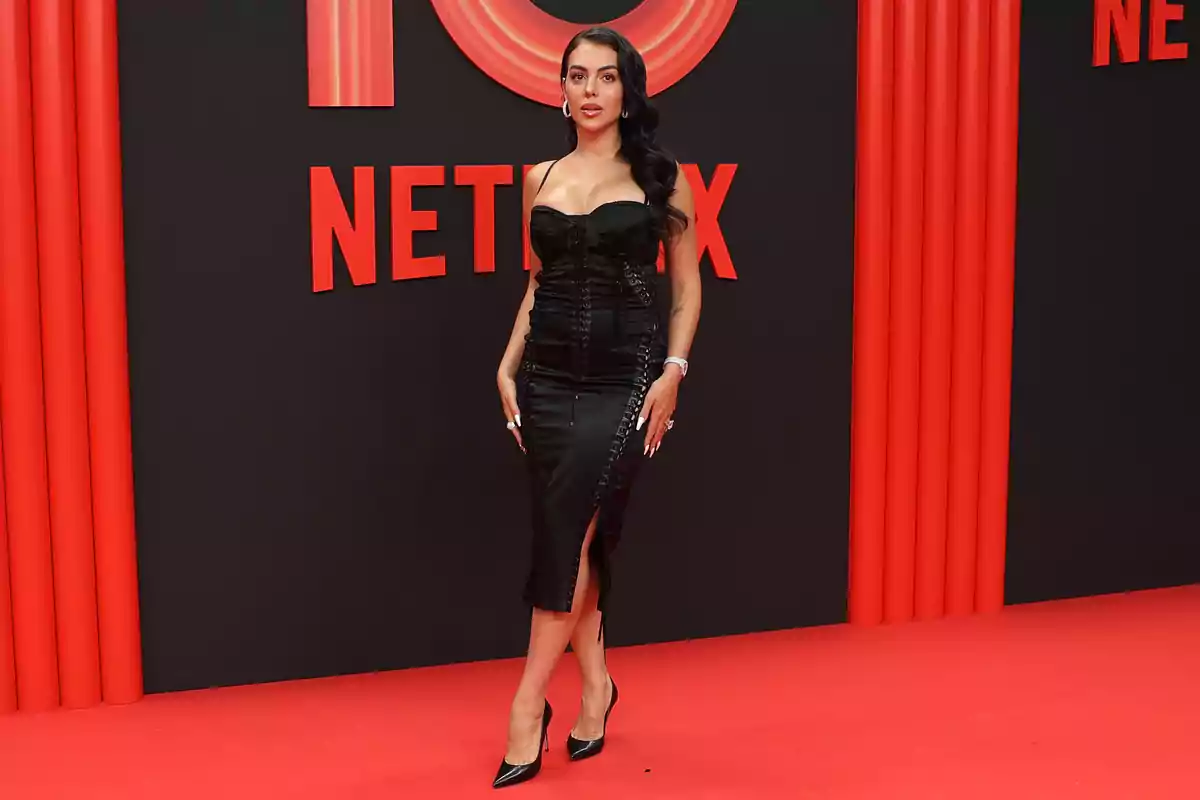 Georgina Rodríguez con vestido negro elegante y tacones posando sobre una alfombra roja con fondo de Netflix Georgina Rodríguez con vestido negro elegante y tacones posando sobre una alfombra roja con fondo de Netflix