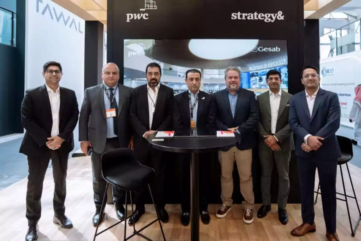 Un grupo de hombres en trajes formales posando juntos frente a un stand de exhibición con logotipos de empresas en el fondo.