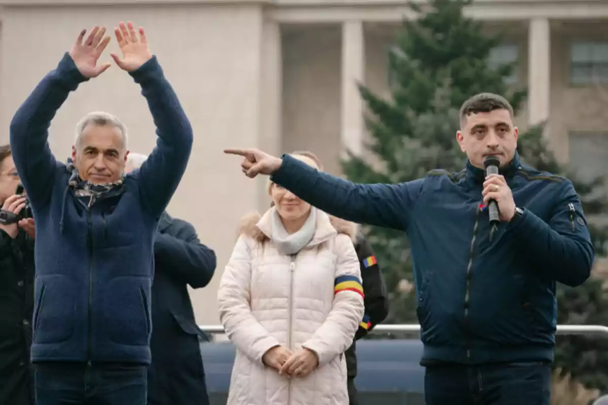 Un hombre con chaqueta azul levanta los brazos mientras otro hombre sostiene un micrófono y señala hacia un lado en un evento al aire libre. Un hombre con chaqueta azul levanta los brazos mientras otro hombre sostiene un micrófono y señala hacia un lado en un evento al aire libre.