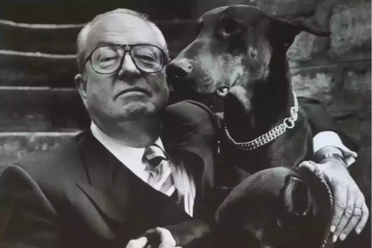 Un hombre mayor con gafas y traje sostiene a un perro grande con collar de cadena en una imagen en blanco y negro.