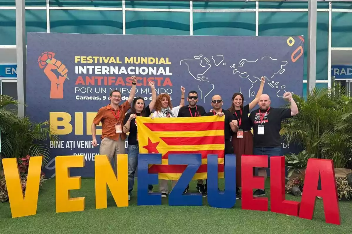 Un grupo de personas posando frente a un cartel del Festival Mundial Internacional Antifascista en Caracas con letras grandes que forman la palabra "Venezuela" y sosteniendo una bandera con franjas amarillas y rojas.