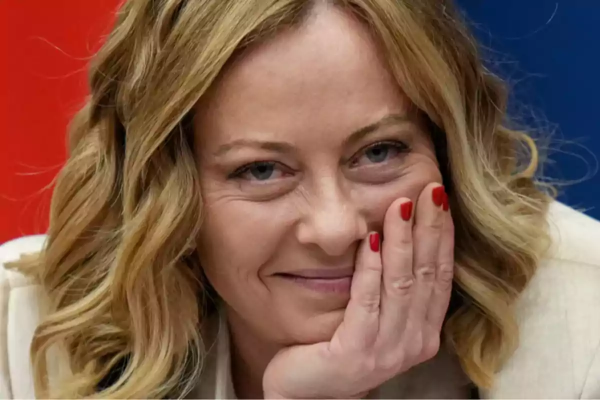 A person with curly blonde hair and red-painted nails smiles while resting their face on their hand.