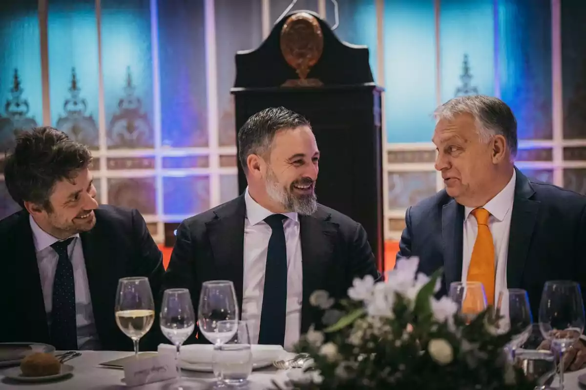 Tres hombres vestidos de traje están sentados en una mesa elegantemente decorada, conversando y sonriendo en un ambiente formal.