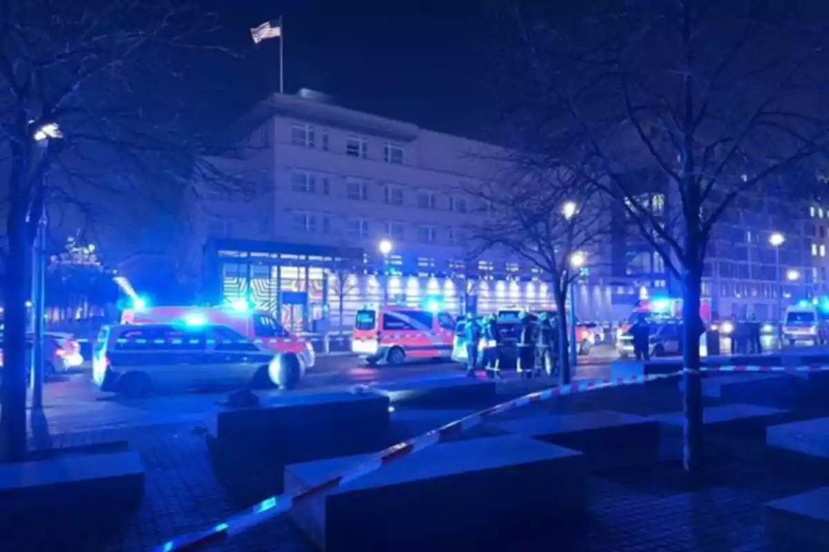 Una escena nocturna muestra un edificio con luces azules de vehículos de emergencia y una bandera ondeando en la parte superior.