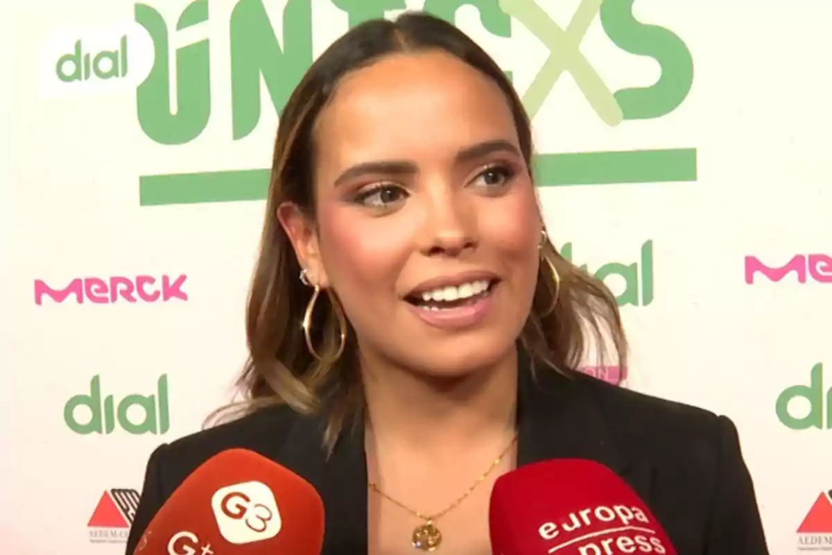 Gloria Camila sonriendo durante una entrevista en un evento con micrófonos de diferentes medios frente a ella y un fondo con logotipos de patrocinadores. Gloria Camila sonriendo durante una entrevista en un evento con micrófonos de diferentes medios frente a ella y un fondo con logotipos de patrocinadores.