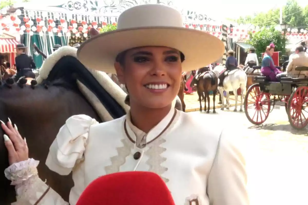Gloria Camila sonriente con sombrero y traje tradicional posa junto a un caballo en un evento al aire libre con carretas y personas vestidas de época. Gloria Camila sonriente con sombrero y traje tradicional posa junto a un caballo en un evento al aire libre con carretas y personas vestidas de época.