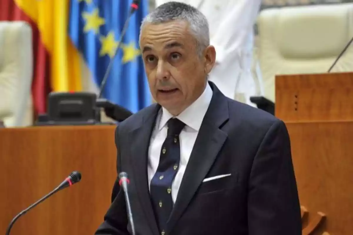 Hombre de cabello canoso y corto hablando en un podio con micrófonos en un entorno institucional con banderas al fondo Hombre de cabello canoso y corto hablando en un podio con micrófonos en un entorno institucional con banderas al fondo