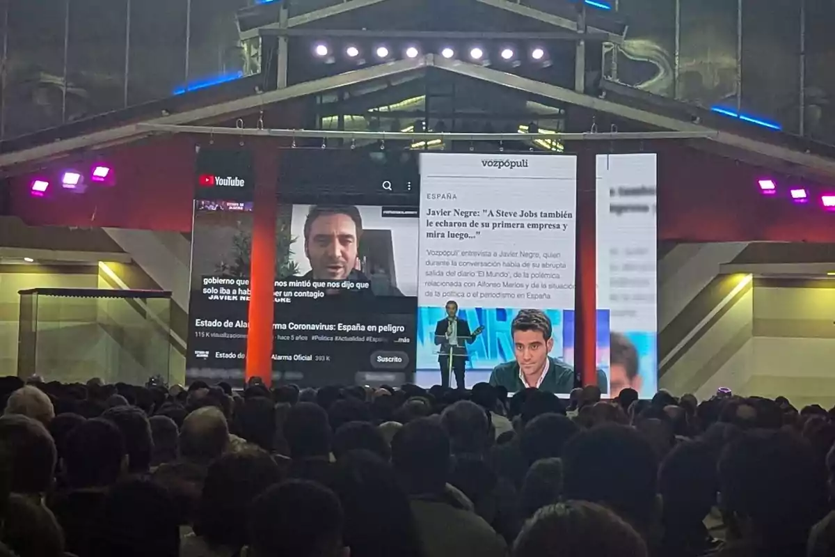 Un grupo de personas observa una presentación en un auditorio, donde se proyectan imágenes y textos en pantallas grandes, incluyendo una entrevista y un video de YouTube. Un grupo de personas observa una presentación en un auditorio, donde se proyectan imágenes y textos en pantallas grandes, incluyendo una entrevista y un video de YouTube.