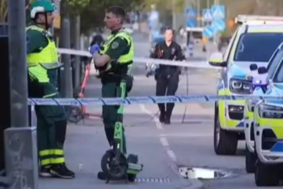 Dos personas con uniformes de emergencia conversan junto a una cinta de seguridad en una calle donde hay vehículos de emergencia estacionados. Dos personas con uniformes de emergencia conversan junto a una cinta de seguridad en una calle donde hay vehículos de emergencia estacionados.
