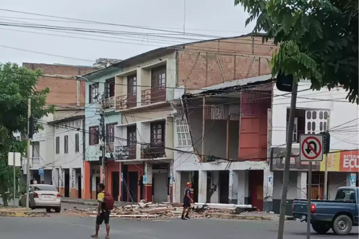 Edificio con fachada dañada y escombros en la acera, con personas caminando cerca y un auto estacionado.