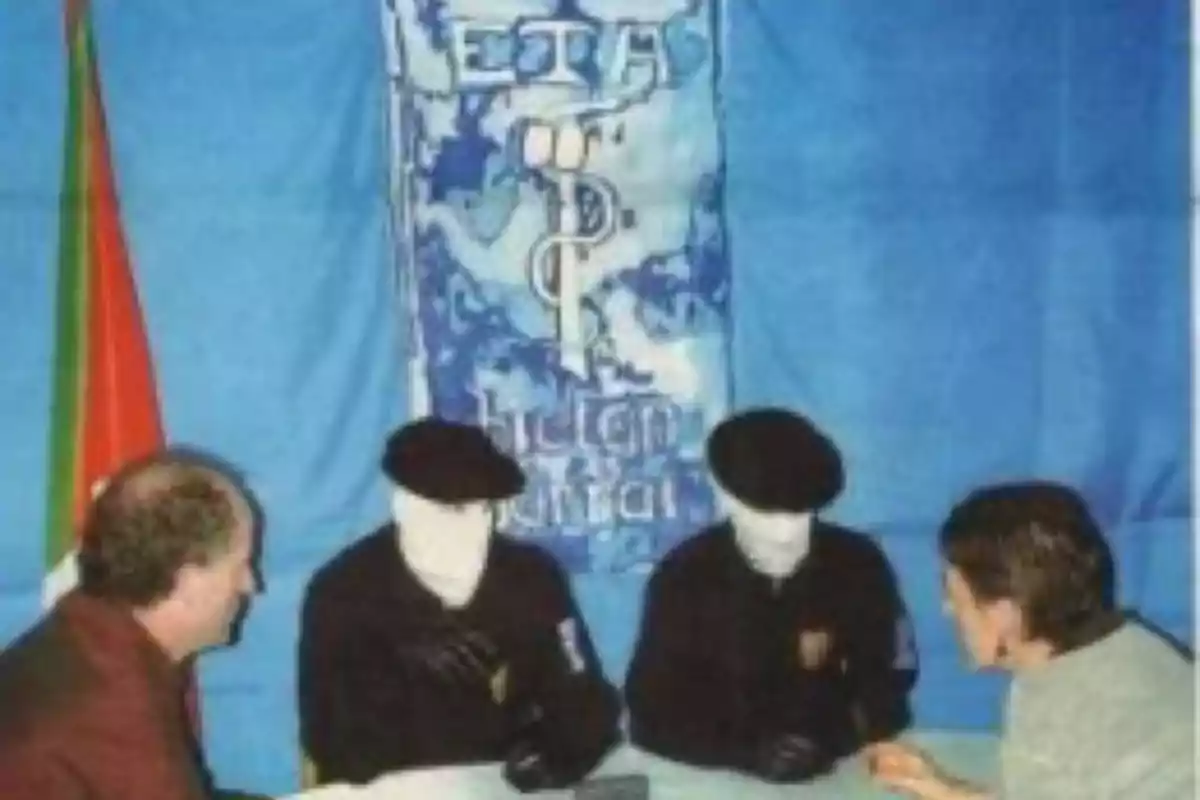 Cuatro personas sentadas en una mesa, dos de ellas con el rostro cubierto, frente a una bandera azul con símbolos y texto. Cuatro personas sentadas en una mesa, dos de ellas con el rostro cubierto, frente a una bandera azul con símbolos y texto.