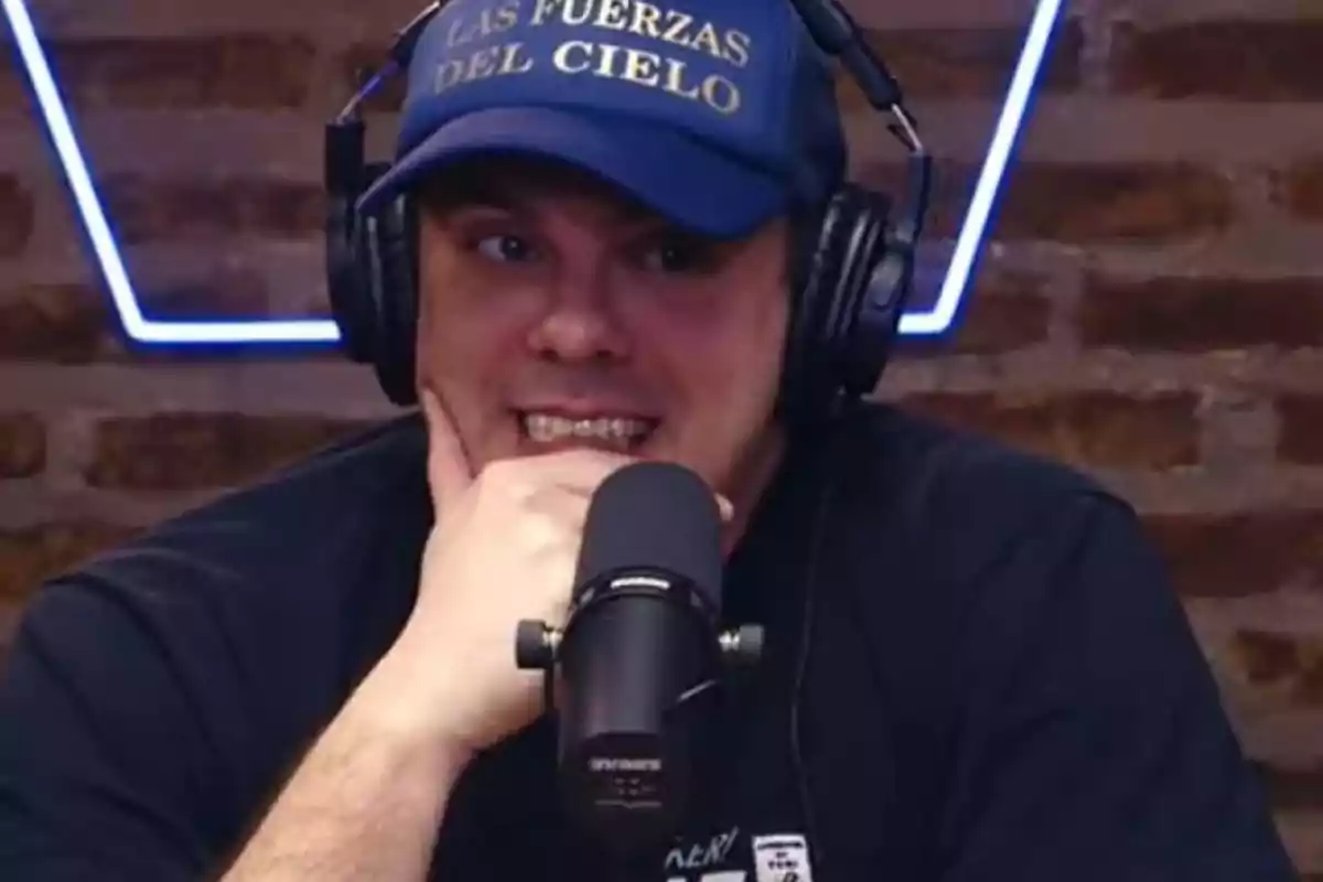 Un hombre con gorra azul y auriculares habla frente a un micrófono en un estudio con paredes de ladrillo. Un hombre con gorra azul y auriculares habla frente a un micrófono en un estudio con paredes de ladrillo.