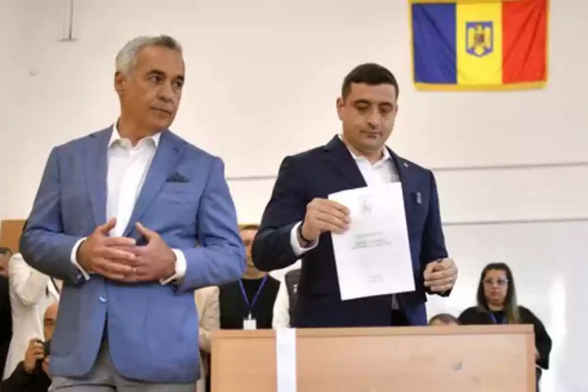 Dos hombres de pie junto a una urna electoral, uno de ellos sostiene un documento, mientras una bandera está colgada en la pared detrás de ellos. Dos hombres de pie junto a una urna electoral, uno de ellos sostiene un documento, mientras una bandera está colgada en la pared detrás de ellos.