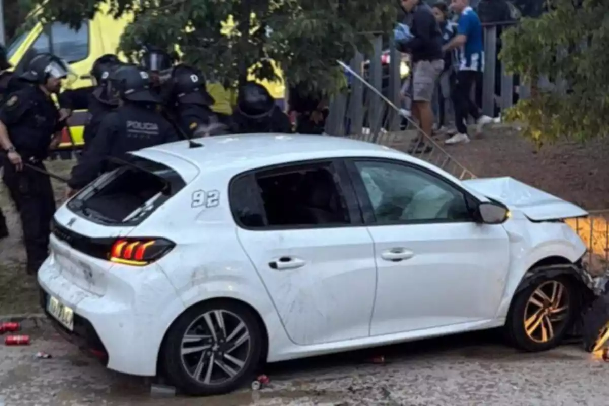 Un coche blanco chocado contra una estructura metálica mientras varios policías con equipo antidisturbios están cerca y algunas personas observan desde el fondo. Un coche blanco chocado contra una estructura metálica mientras varios policías con equipo antidisturbios están cerca y algunas personas observan desde el fondo.