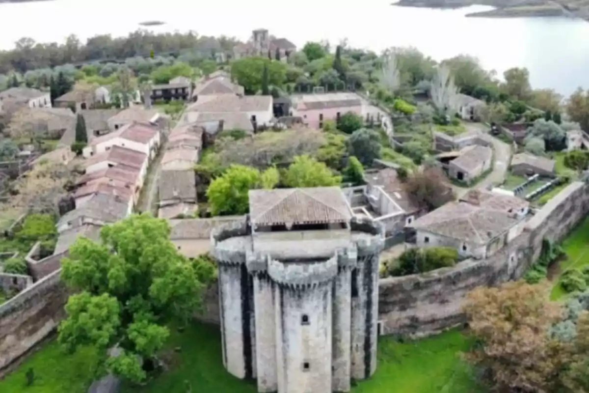 Vista aérea de un antiguo castillo rodeado de un pequeño pueblo con casas de techos de tejas y vegetación abundante, ubicado junto a un cuerpo de agua.