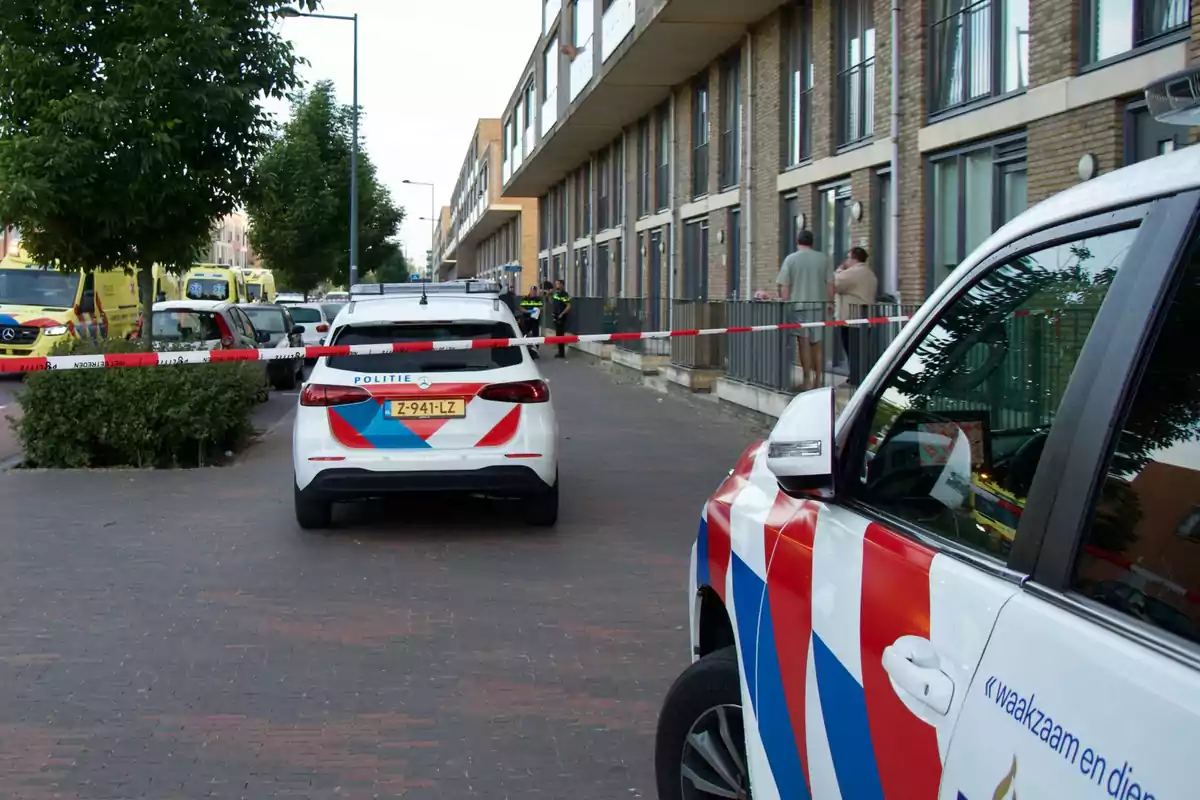 Una escena urbana con vehículos de policía y ambulancias estacionados en una calle, delimitada por cinta de seguridad, mientras algunas personas observan desde la acera. Una escena urbana con vehículos de policía y ambulancias estacionados en una calle, delimitada por cinta de seguridad, mientras algunas personas observan desde la acera.