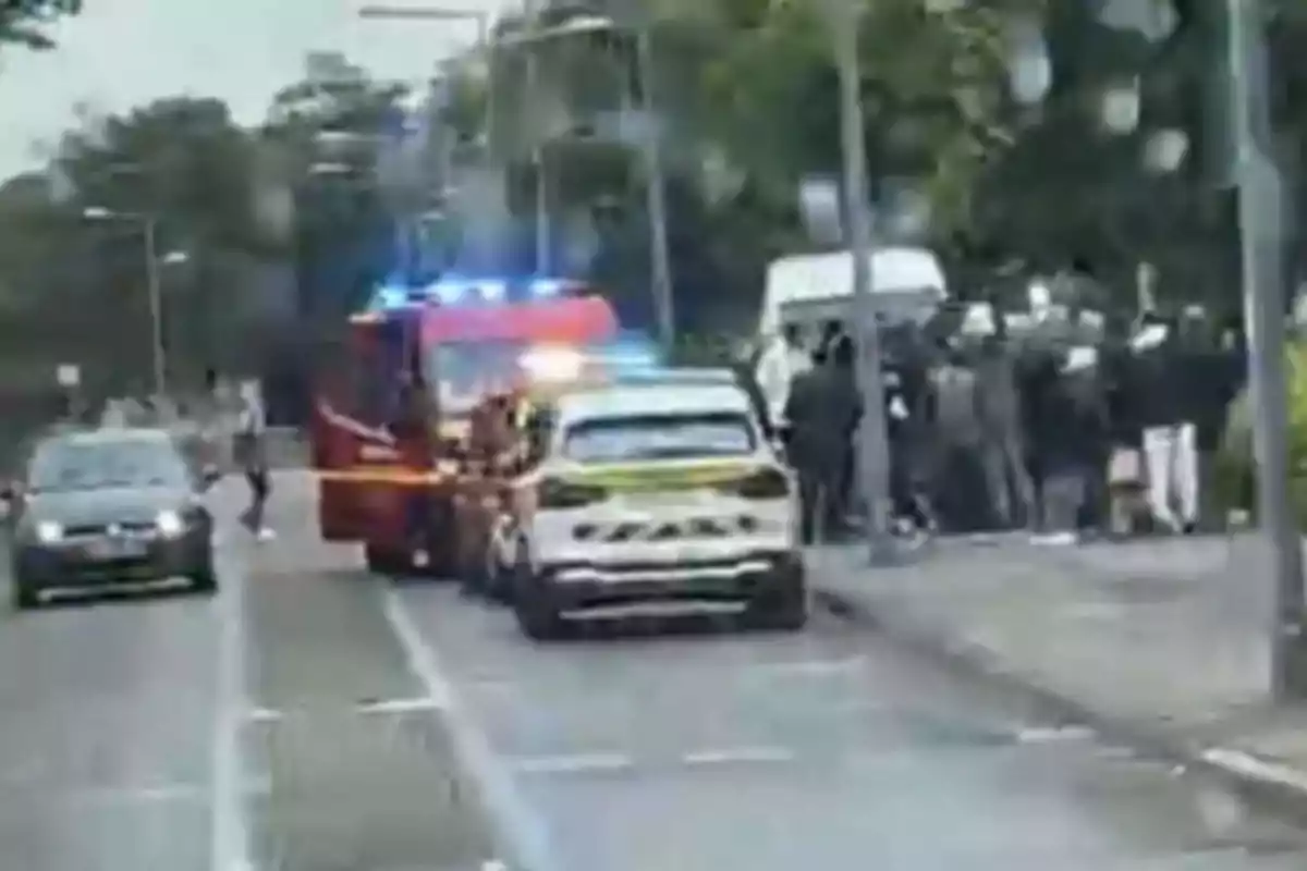 Un coche de policía y un camión de bomberos con luces encendidas están estacionados en una calle junto a un grupo de personas.
