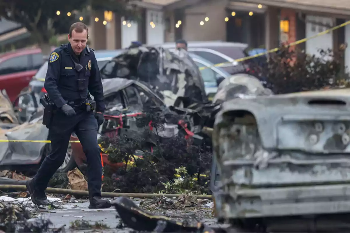 Un oficial de policía camina entre los restos de un accidente con vehículos quemados y cinta de seguridad en una calle residencial. Un oficial de policía camina entre los restos de un accidente con vehículos quemados y cinta de seguridad en una calle residencial.