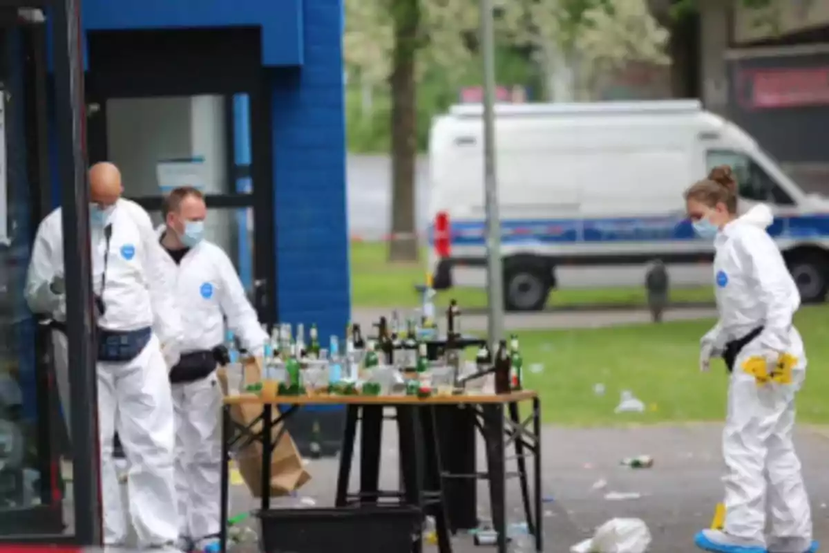 Personas con trajes de protección y mascarillas inspeccionan una mesa llena de botellas vacías al aire libre, con una furgoneta blanca en el fondo. Personas con trajes de protección y mascarillas inspeccionan una mesa llena de botellas vacías al aire libre, con una furgoneta blanca en el fondo.