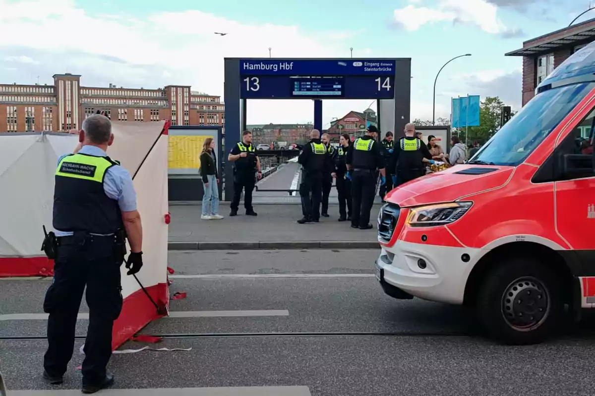 Un grupo de policías y personal de seguridad se encuentra en una estación de tren en Hamburgo, con una ambulancia roja estacionada cerca y una estructura blanca y roja en el lugar. Un grupo de policías y personal de seguridad se encuentra en una estación de tren en Hamburgo, con una ambulancia roja estacionada cerca y una estructura blanca y roja en el lugar.