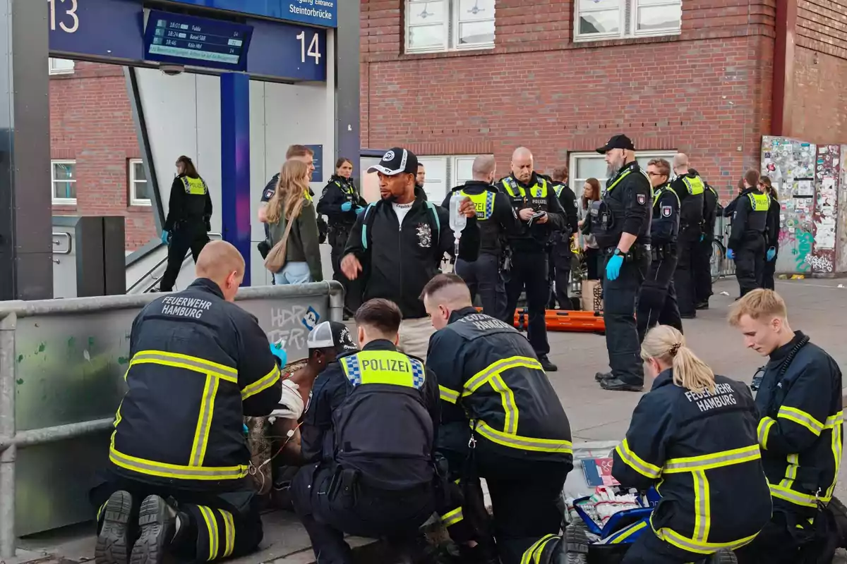 Un grupo de policías y bomberos atiende una situación en una estación de tren, mientras varias personas observan.