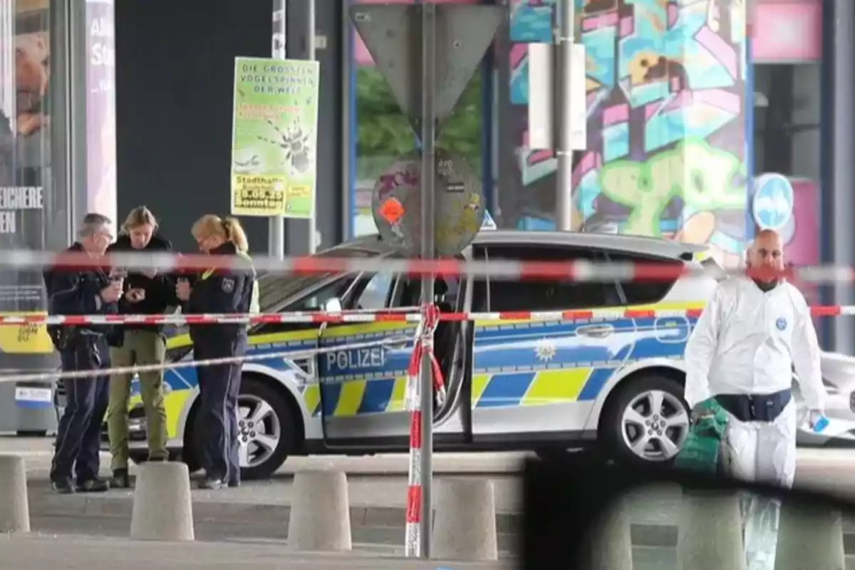 Un grupo de policías y un investigador forense trabajan en una escena acordonada con un coche patrulla en un entorno urbano.