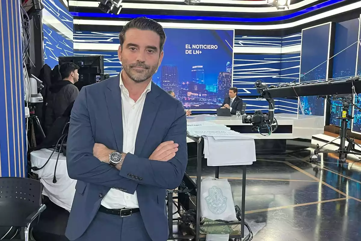 Un hombre de traje azul y camisa blanca está de pie con los brazos cruzados en un estudio de televisión, con un set de noticias y equipo de grabación en el fondo.