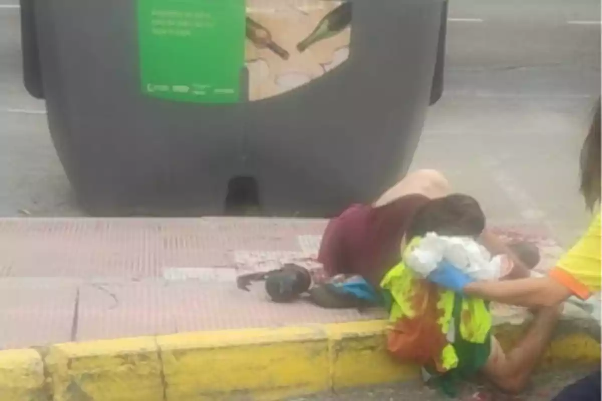 Persona herida recostada en la acera junto a un contenedor de reciclaje mientras otra persona le brinda primeros auxilios Persona herida recostada en la acera junto a un contenedor de reciclaje mientras otra persona le brinda primeros auxilios
