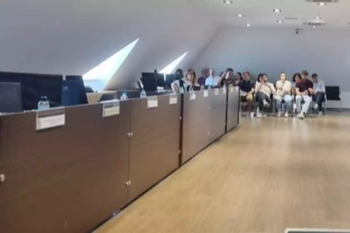 Personas sentadas al fondo de una sala de reuniones con escritorios y sillas vacías en primer plano Personas sentadas al fondo de una sala de reuniones con escritorios y sillas vacías en primer plano
