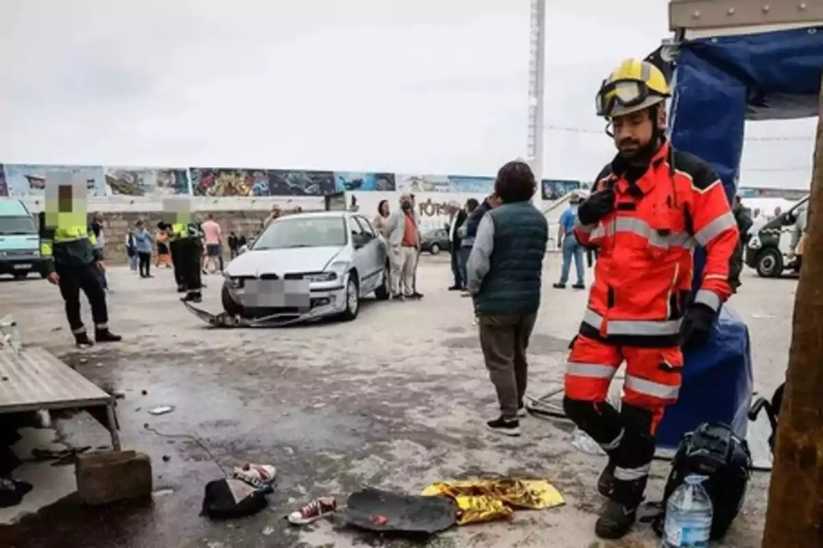 Un bombero con uniforme naranja y casco amarillo está de pie junto a una zona donde ocurrió un accidente de tráfico, con un coche dañado y varias personas y policías alrededor, mientras objetos personales y equipo de emergencia están en el suelo.