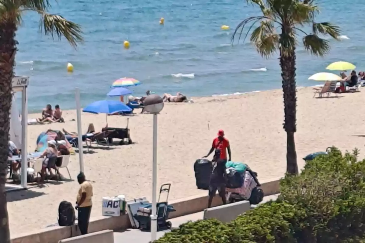 Personas en la playa disfrutando del sol y el mar, con sombrillas de colores y dos vendedores ambulantes empujando carros llenos de mercancía por el paseo junto a la arena Personas en la playa disfrutando del sol y el mar, con sombrillas de colores y dos vendedores ambulantes empujando carros llenos de mercancía por el paseo junto a la arena