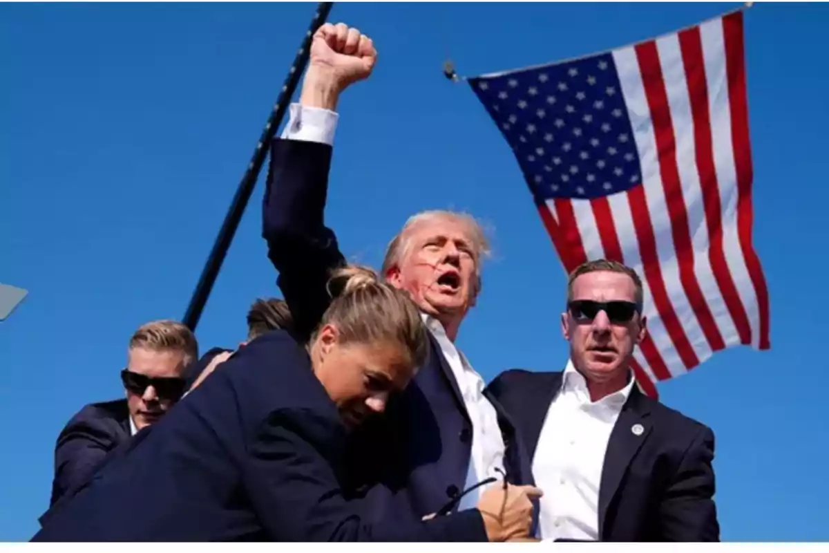 Un grupo de personas, algunas con gafas de sol, rodea a un hombre que levanta el puño, con una bandera de Estados Unidos ondeando en el fondo.