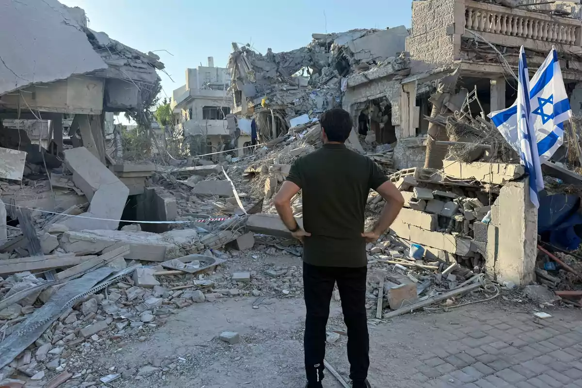 Un hombre de espaldas observa los escombros de varios edificios destruidos, entre los restos hay dos banderas de Israel ondeando Un hombre de espaldas observa los escombros de varios edificios destruidos, entre los restos hay dos banderas de Israel ondeando