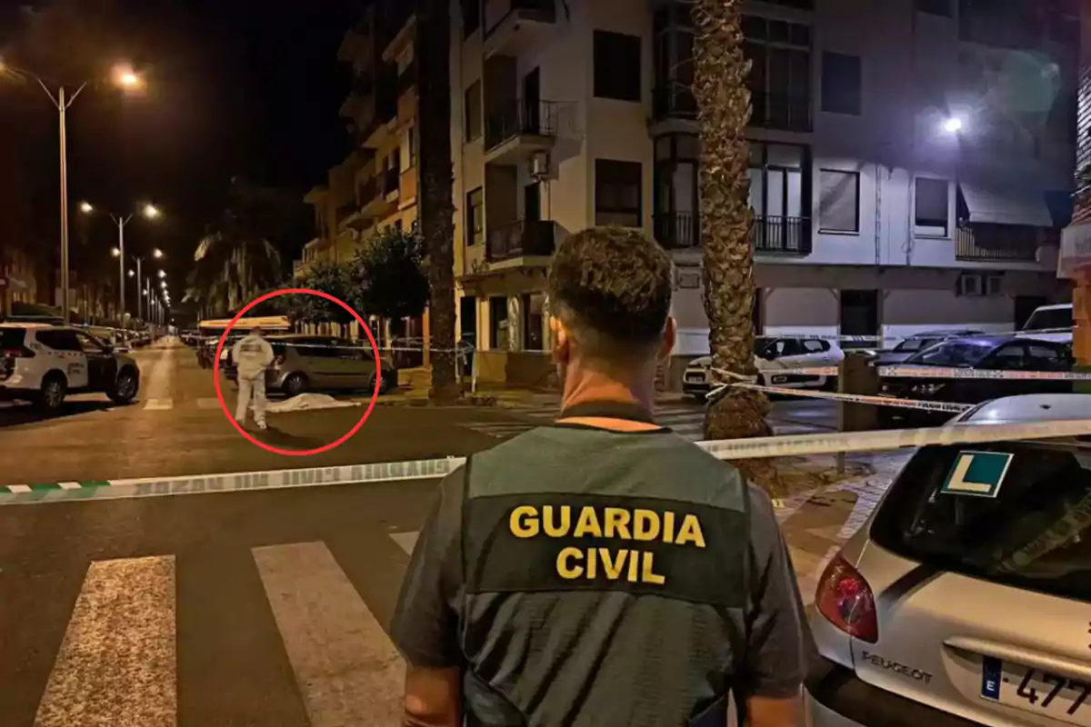 Un agente de la Guardia Civil observa una escena acordonada en una calle durante la noche, con un investigador forense trabajando en el lugar.