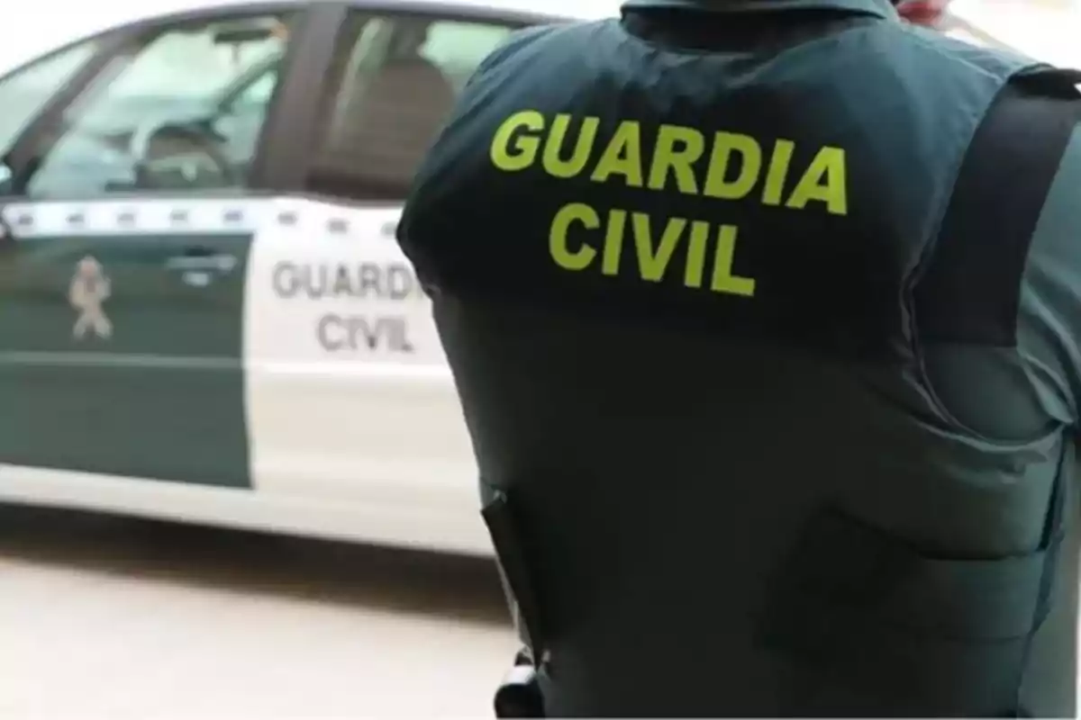 Agente de la Guardia Civil de espaldas con un coche patrulla al fondo.