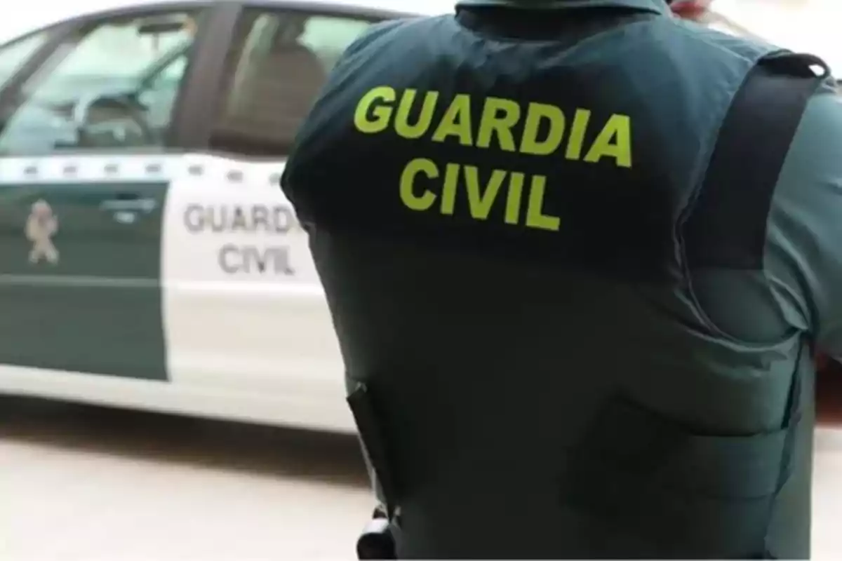 Un agente de la Guardia Civil de espaldas con un coche patrulla al fondo.