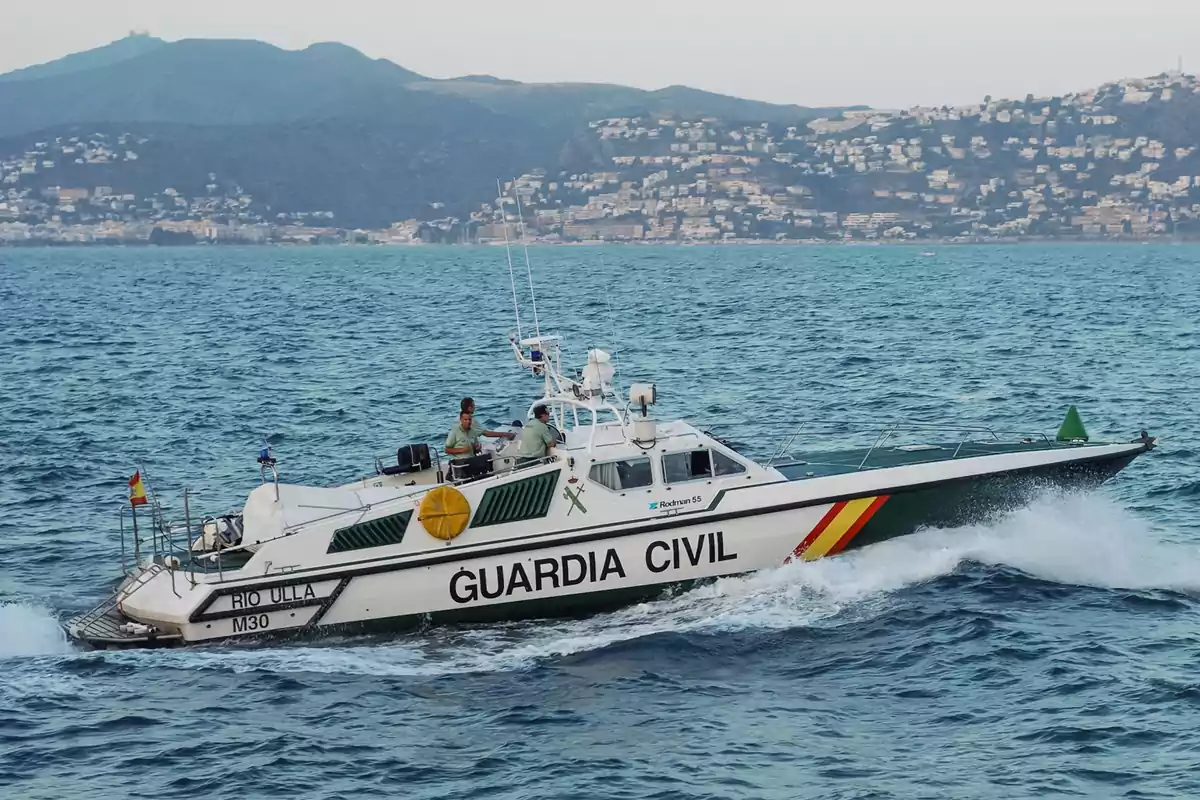Una lancha de la Guardia Civil navegando en el mar con un paisaje montañoso y urbano al fondo. Una lancha de la Guardia Civil navegando en el mar con un paisaje montañoso y urbano al fondo.