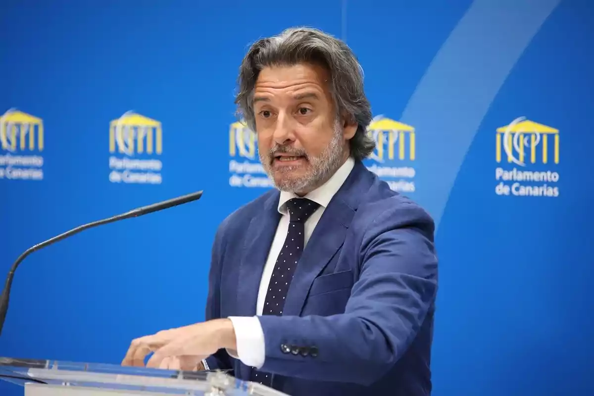 Un hombre con traje azul y corbata habla en un podio frente a un fondo azul con el logo del Parlamento de Canarias.