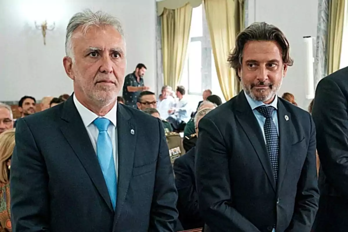 Dos hombres de pie en un evento formal, ambos con trajes oscuros y corbatas, rodeados de otras personas sentadas en un salón con cortinas amarillas. Dos hombres de pie en un evento formal, ambos con trajes oscuros y corbatas, rodeados de otras personas sentadas en un salón con cortinas amarillas.