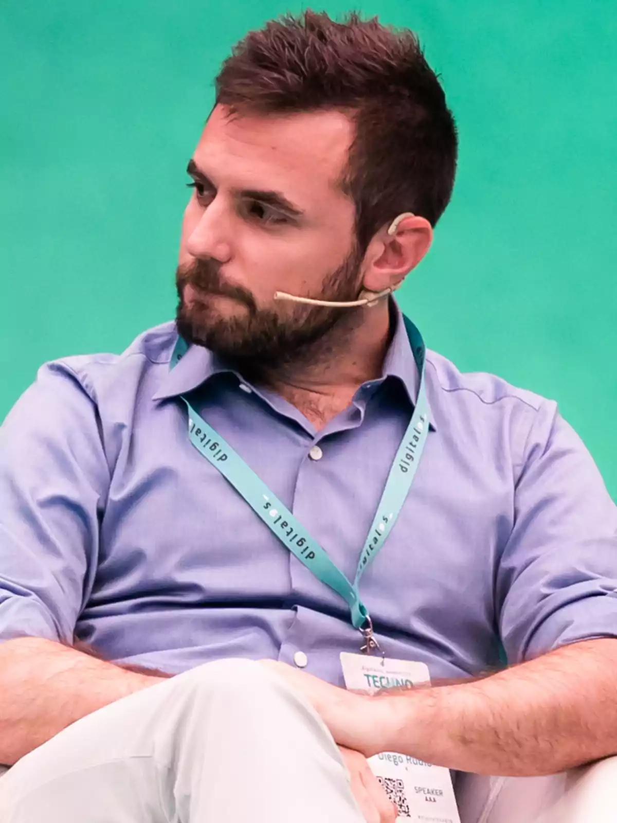 Hombre con barba y cabello corto, usando una camisa azul y un micrófono de diadema, sentado con los brazos cruzados y una credencial colgada al cuello, sobre un fondo verde. Hombre con barba y cabello corto, usando una camisa azul y un micrófono de diadema, sentado con los brazos cruzados y una credencial colgada al cuello, sobre un fondo verde.
