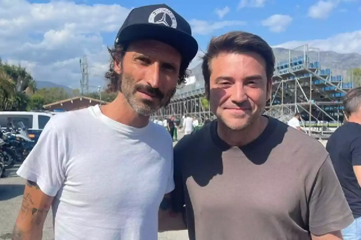 Dos hombres posando juntos al aire libre con una estructura metálica y montañas al fondo.