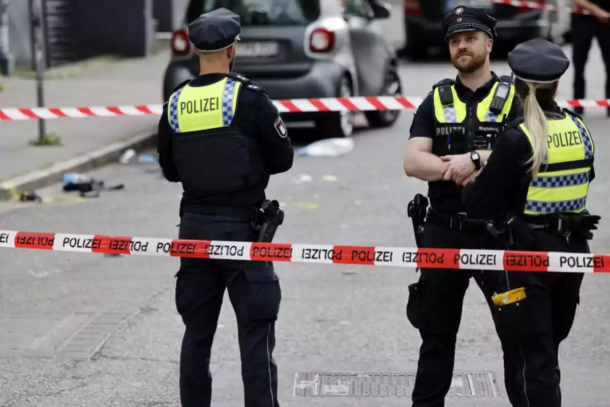 Tres policías alemanes con chalecos amarillos y uniformes oscuros resguardan una zona acordonada con cinta roja y blanca que dice Polizei en una calle urbana