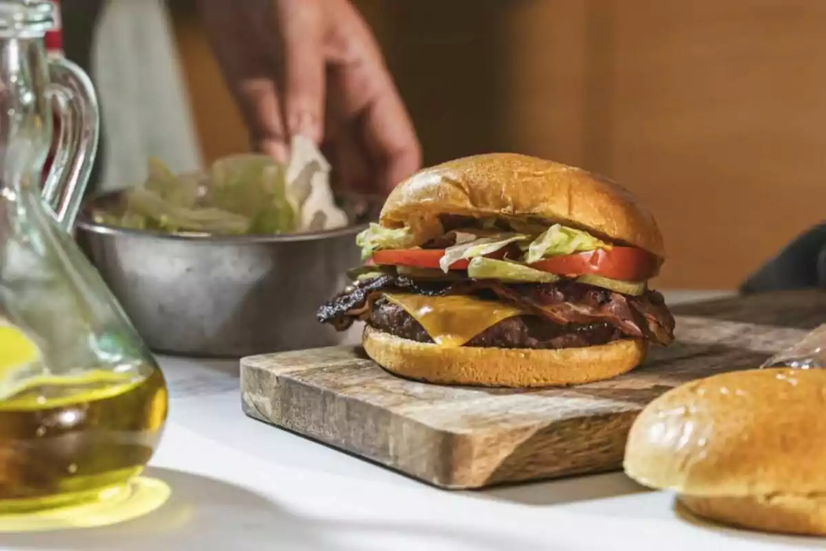 Hamburguesa con queso, tocino, lechuga y jitomate sobre una tabla de madera con una mano al fondo preparando ingredientes
