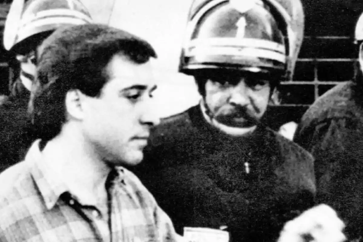 Un hombre joven con camisa a cuadros es escoltado por un policía con casco en una imagen en blanco y negro Un hombre joven con camisa a cuadros es escoltado por un policía con casco en una imagen en blanco y negro