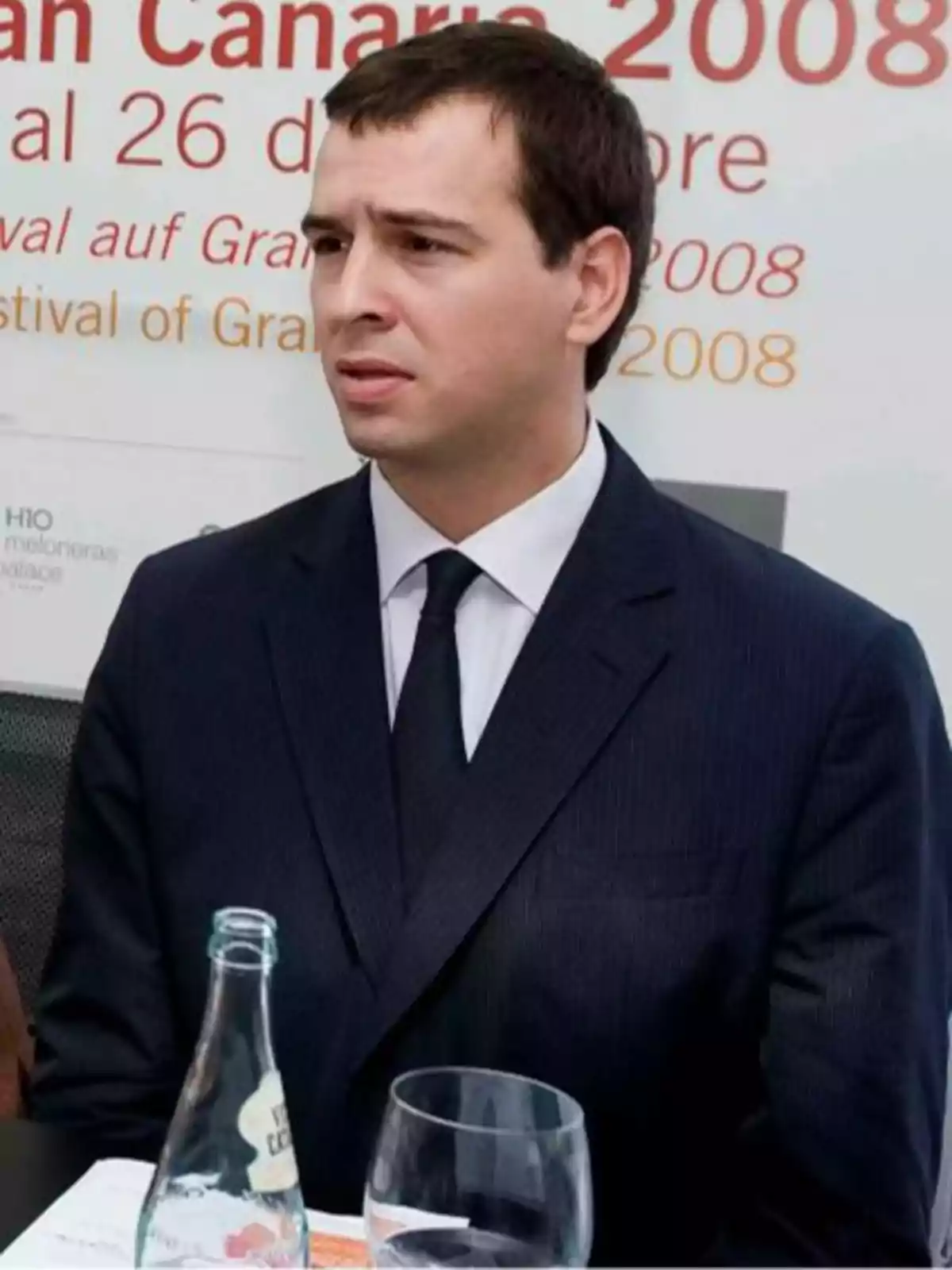 A man in a dark suit and tie sits at a table with a bottle of water and a glass, with a poster in the background mentioning an event in Gran Canaria in 2008. A man in a dark suit and tie sits at a table with a bottle of water and a glass, with a poster in the background mentioning an event in Gran Canaria in 2008.