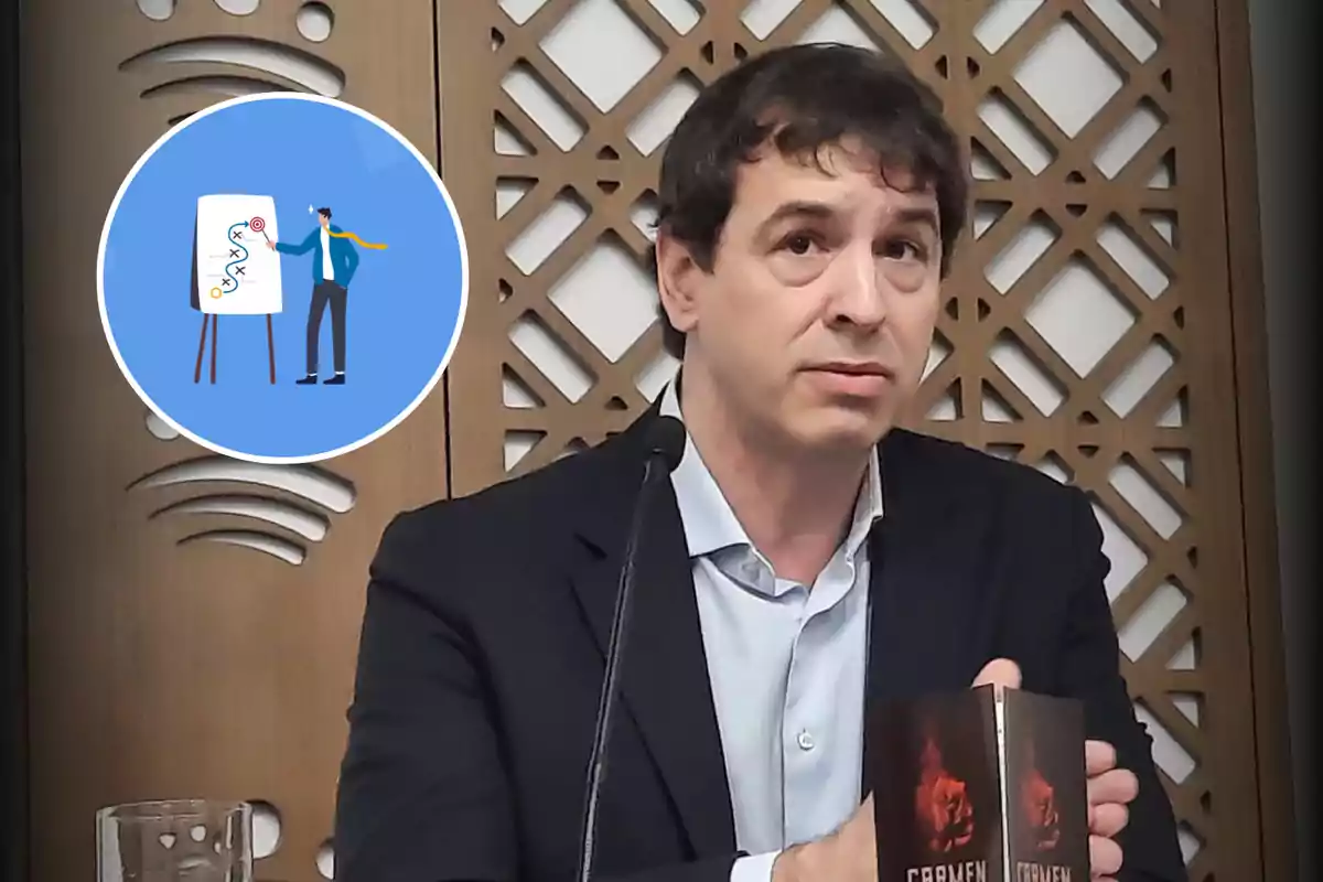 Un hombre en una conferencia sostiene un libro con una portada oscura mientras está sentado frente a un fondo decorativo de madera.