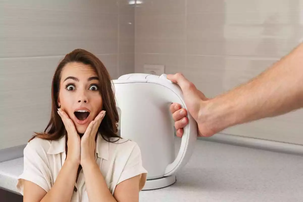 Una mujer con expresión de sorpresa junto a una mano que sostiene una tetera eléctrica en una cocina.