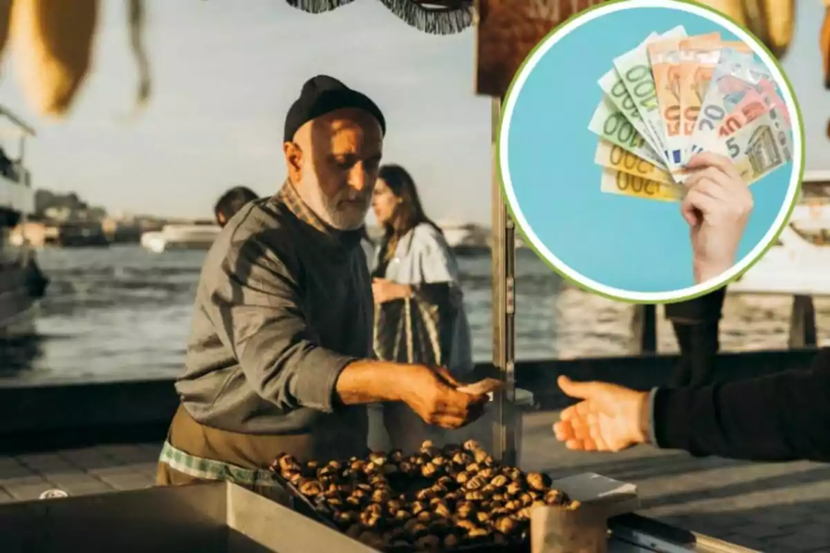 Un hombre mayor vende castañas asadas en un puesto al aire libre junto a un río, mientras entrega dinero a un cliente; en la esquina superior derecha de la imagen hay un círculo que muestra una mano sosteniendo varios billetes de euro.